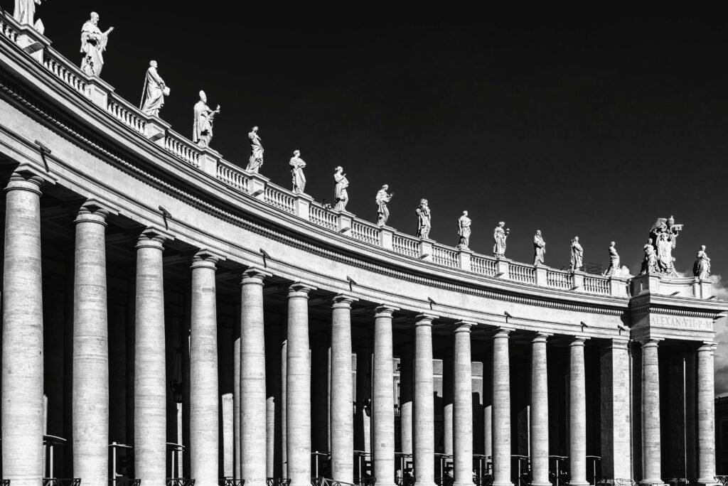 Dramatic black and white photograph of Vatican colonnade with statues, highlighting classic architecture under a clear sky.