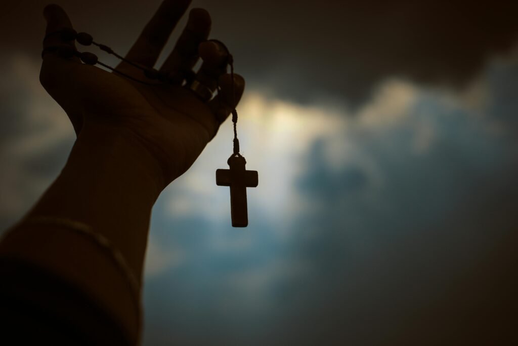 Silhouette of a hand holding a rosary with a cross against a dramatic sky, symbolizing faith and spirituality.