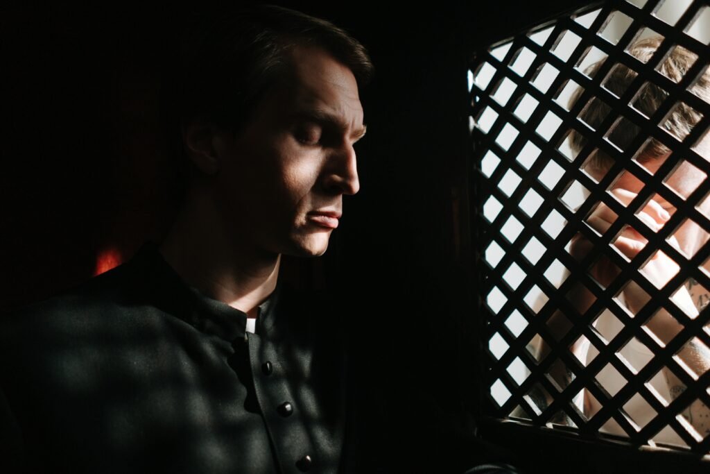 A priest listens to a penitent in a dimly lit confessional, embodying themes of faith and repentance.